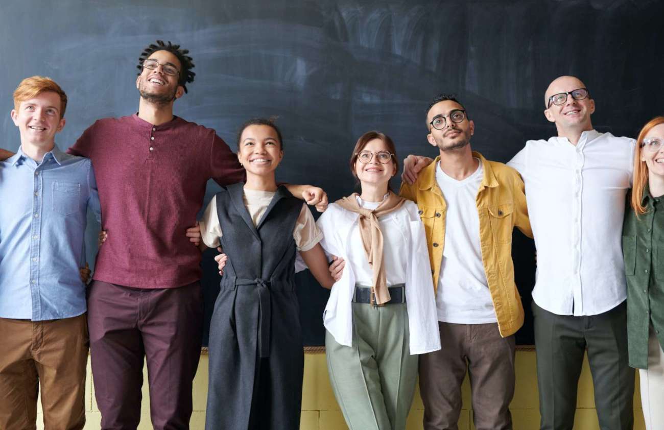 7 smiling workers linking arms behind a blackboard in a happy workplace